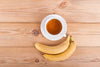 Two bananas sitting beside tea cup on wooden table.